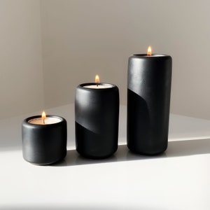 May include: Three black candle holders with lit candles. The candle holders are of different sizes and are arranged in a row from smallest to largest.