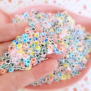 May include: A bowl filled with colorful paw print shaped resin embellishments. The paw prints are in various colors including pink, blue, green, yellow, and black.