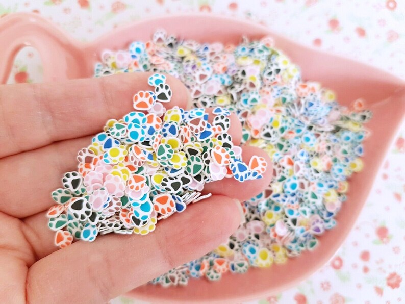 May include: A bowl filled with colorful paw print shaped resin embellishments. The paw prints are in various colors including pink, blue, green, yellow, and black.