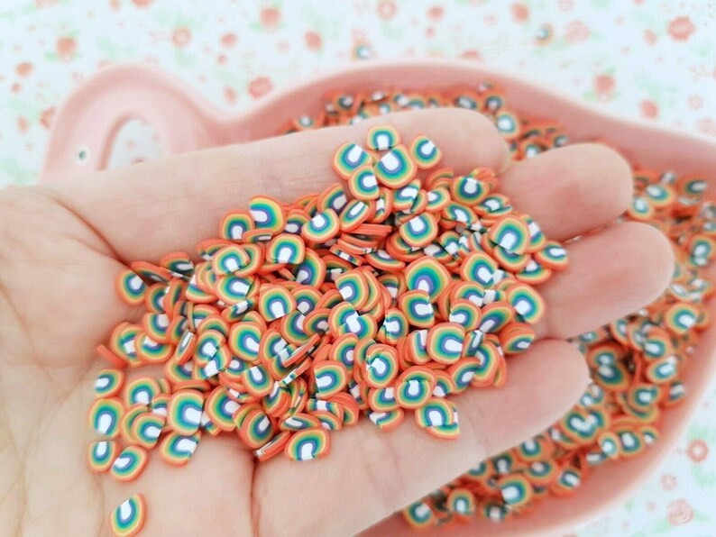 May include: A large pile of rainbow-colored, flat, round, polymer clay sprinkles. The sprinkles are shaped like half circles with a white center and rainbow stripes.