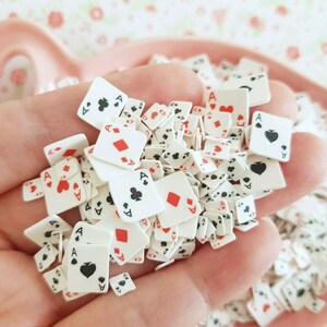 May include: A hand holding a pile of small white playing card charms with red and black details. The charms are shaped like playing cards and feature the ace of spades, hearts, diamonds, and clubs.