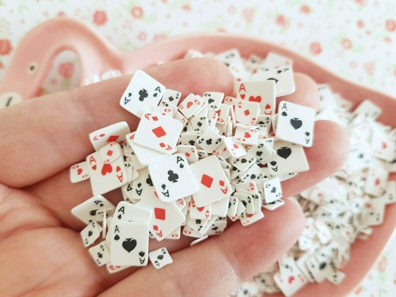 May include: A hand holding a pile of small white playing card charms with red and black details. The charms are shaped like playing cards and feature the ace of spades, hearts, diamonds, and clubs.