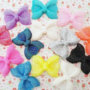 Può includere: Assortimento di 12 fiocchi di strass in vari colori, tra cui rosa, blu, bianco, nero e giallo. I fiocchi sono realizzati in un materiale lucido e hanno una finitura scintillante.