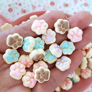 May include: A collection of small, colourful paw print charms in various pastel colours, including pink, blue, and brown. The charms are made of polymer clay and are perfect for adding a touch of whimsy to jewellery, crafts, and other projects.