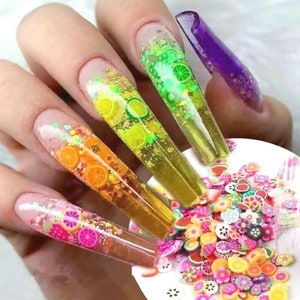 May include: Close-up of five long, clear acrylic nails with colorful glitter and fruit designs. The nails are decorated with small, colorful fruit charms and glitter. The nails are on a white surface with a scattering of colorful fruit charms.