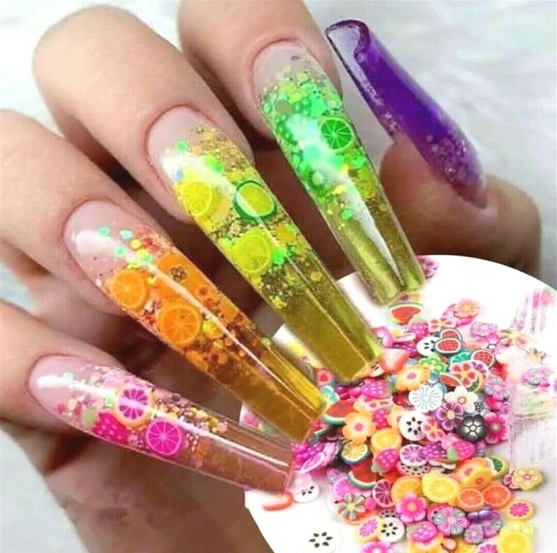 May include: Close-up of five long, clear acrylic nails with colorful glitter and fruit designs. The nails are decorated with small, colorful fruit charms and glitter. The nails are on a white surface with a scattering of colorful fruit charms.