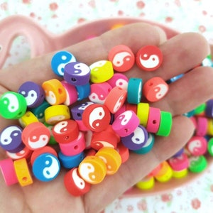 Puede incluir: Una colección de cuentas redondas de colores con un símbolo yin yang en cada cuenta. Las cuentas son de plástico y tienen un agujero en el centro para enhebrar.