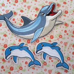May include: Three embroidered iron-on patches of dolphins. The largest dolphin is blue and grey with an open mouth. The two smaller dolphins are blue and white.
