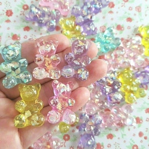 May include: A selection of colourful, translucent gummy bear-shaped charms. The charms are in various colours, including pink, yellow, blue, and purple, with iridescent glitter embedded within. The charms are held in a hand, with more scattered in the background.