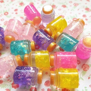 May include: A collection of small glass bottles filled with colourful glitter and iridescent flakes. The bottles have gold lids and are arranged on a pink and white floral patterned surface.