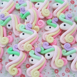 Könnte beinhalten: Eine Sammlung von pastellfarbenen Einhornkopf-Anhängern aus Polymer Clay. Die Anhänger zeigen einen weißen Einhornkopf mit einer Regenbogenmähne und einem Regenbogen-Schwanz.