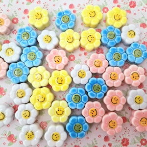 Op de afbeelding: Een verzameling keramische knopen in de vorm van bloemen met smiley-gezichten. De knopen zijn in verschillende kleuren, waaronder geel, blauw, roze en wit.