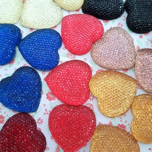 Op de afbeelding: Een verzameling hartvormige versieringen in verschillende kleuren, waaronder rood, blauw, goud en zwart. Elk hart is bedekt met kleine, glinsterende strass steentjes. De harten zijn gerangschikt op een bloemmotief achtergrond.