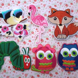 May include: A set of seven embroidered iron-on patches featuring various animals and designs. The patches include a bluebird on a branch with pink flowers, a pink flamingo, a red fox, a green caterpillar, a pink owl, and a blue owl.