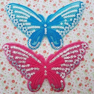 Puede incluir: Dos apliques de mariposa con lentejuelas, uno azul y otro rosa, con lentejuelas plateadas. Las mariposas están sobre un fondo con estampado floral.