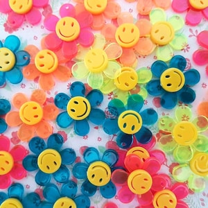 May include: Assortment of colourful plastic flower-shaped buttons with smiley faces. The buttons are in shades of yellow, orange, pink, blue, and green.