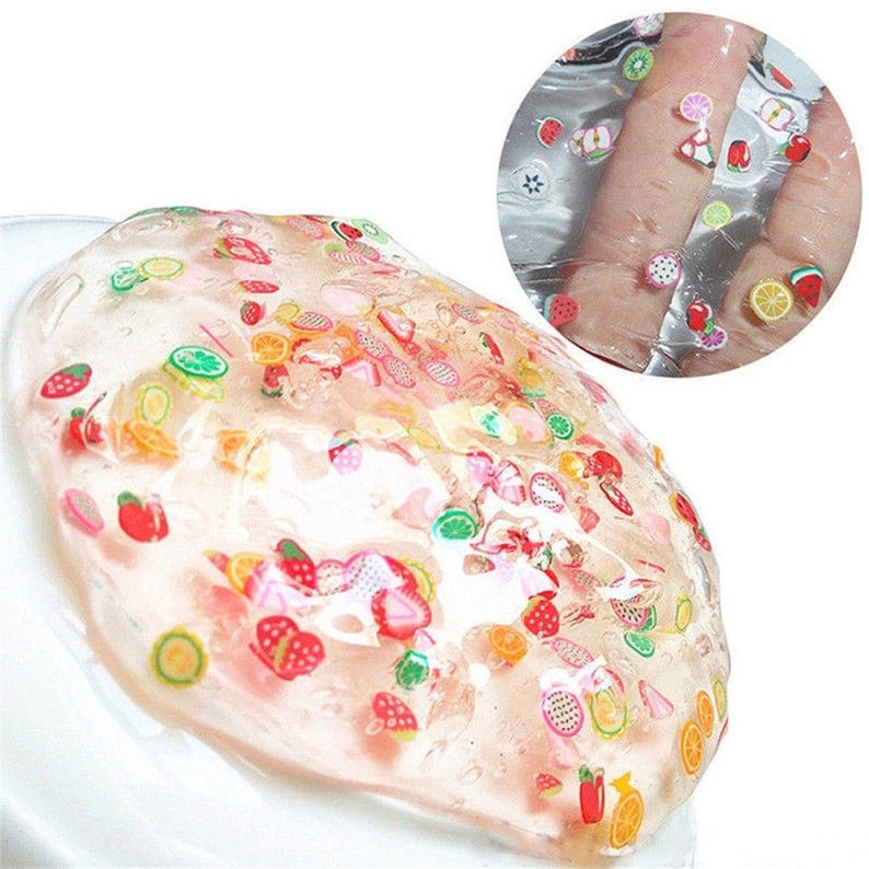 May include: Clear slime with a variety of colorful fruit and flower shaped charms. The slime is spread out on a white surface.