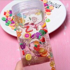 May include: A clear glass jar filled with a clear liquid and colorful fruit-shaped charms. The charms are in various colors, including red, yellow, green, and purple. The jar has a white lid.