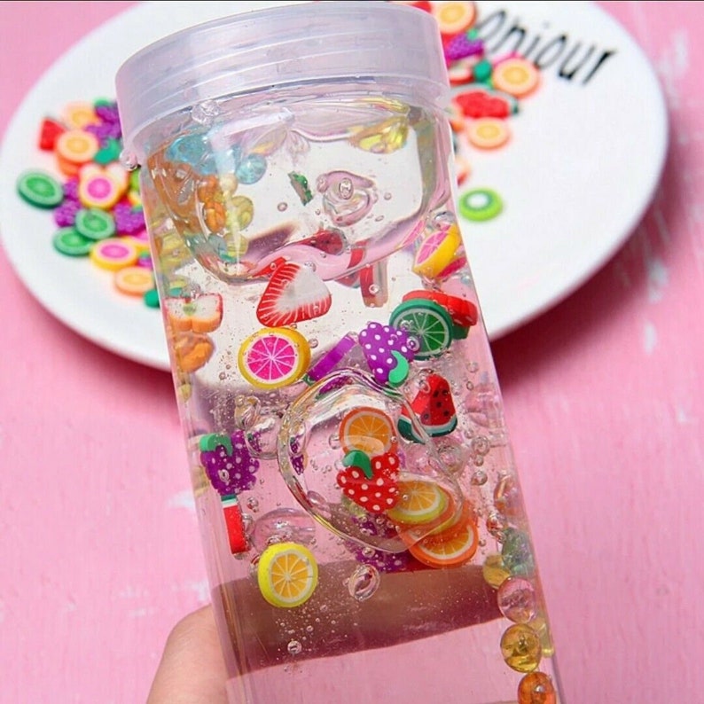 May include: A clear glass jar filled with a clear liquid and colorful fruit-shaped charms. The charms are in various colors, including red, yellow, green, and purple. The jar has a white lid.