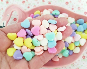 May include: A collection of pastel-colored heart-shaped beads in shades of yellow, pink, blue, purple, and white. The beads are held in a pink flamingo-shaped dish, perfect for crafting or jewelry making.