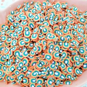 May include: A bowl filled with rainbow-colored polymer clay sprinkles. The sprinkles are shaped like half circles with a rainbow design.