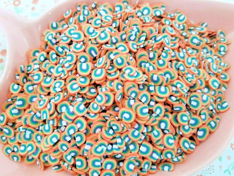 May include: A bowl filled with rainbow-colored polymer clay sprinkles. The sprinkles are shaped like half circles with a rainbow design.