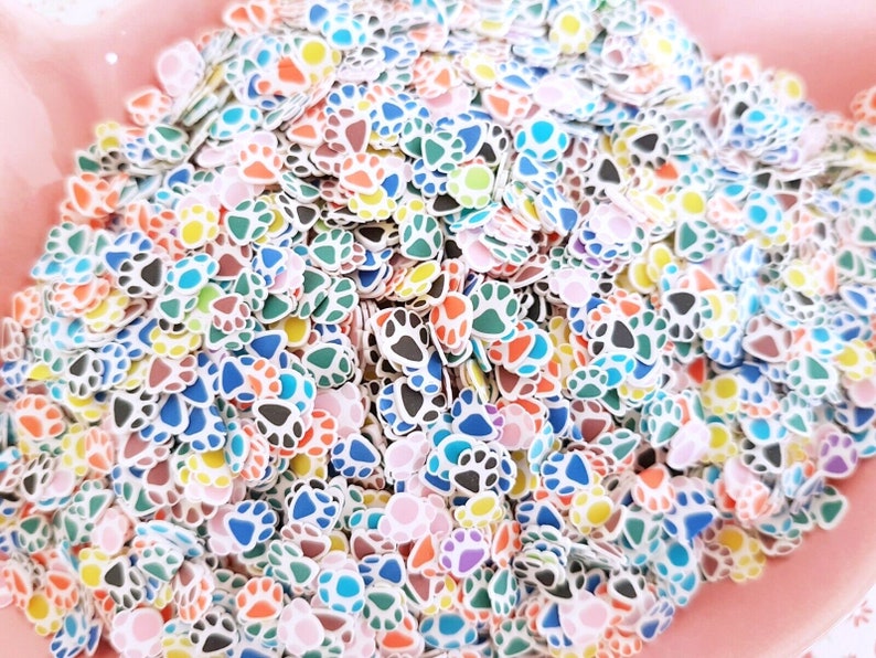 May include: A large pile of colorful paw print shaped sprinkles. The sprinkles are white with different colored accents, including blue, green, pink, orange, yellow, and brown.