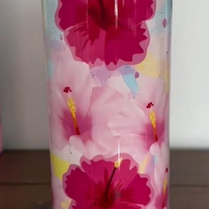 Op de afbeelding: Een hoge, cilindrische beker met een levendig bloemenontwerp. De beker heeft grote, dieproze hibiscusbloemen tegen een achtergrond van lichtere roze en pastelkleurige aquarelspatten. De beker wordt gepresenteerd op een witte sokkel.
