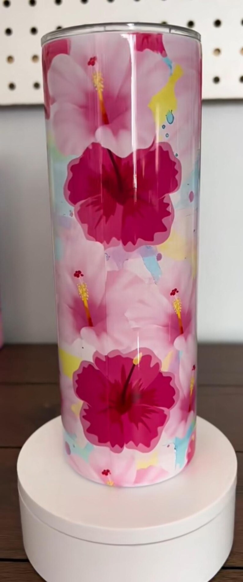 Op de afbeelding: Een hoge, cilindrische beker met een levendig bloemenontwerp. De beker heeft grote, dieproze hibiscusbloemen tegen een achtergrond van lichtere roze en pastelkleurige aquarelspatten. De beker wordt gepresenteerd op een witte sokkel.
