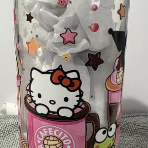 Pu&ograve; includere: Un barattolo di vetro trasparente con un coperchio in bamb&ugrave; con un design di Hello Kitty. Il barattolo ha una tazza rosa di Hello Kitty con il testo "Cafecito y Chismes". Il barattolo presenta anche un personaggio di rana verde e altri dettagli colorati.