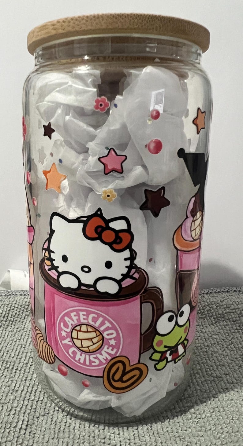 Pu&ograve; includere: Un barattolo di vetro trasparente con un coperchio in bamb&ugrave; con un design di Hello Kitty. Il barattolo ha una tazza rosa di Hello Kitty con il testo "Cafecito y Chismes". Il barattolo presenta anche un personaggio di rana verde e altri dettagli colorati.