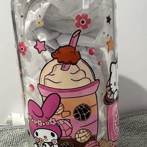 Pu&ograve; includere: Un barattolo di vetro trasparente con un coperchio di legno presenta un design colorato con un coniglio rosa e bianco, un personaggio di Hello Kitty e un bubble tea rosa e arancione con topping al cioccolato marrone. Il design include anche fiori rosa e gialli, stelle e pasticcini marroni e bianchi.