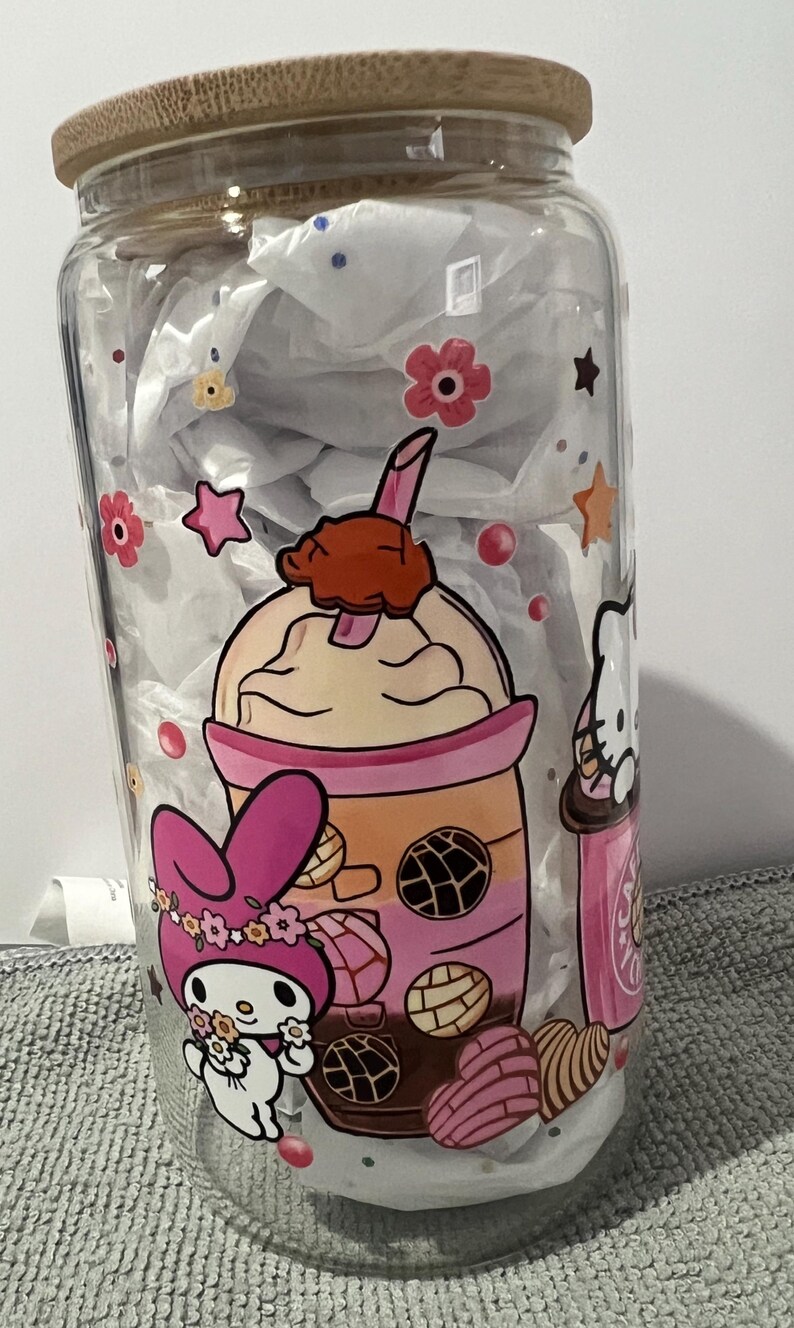 Pu&ograve; includere: Un barattolo di vetro trasparente con un coperchio di legno presenta un design colorato con un coniglio rosa e bianco, un personaggio di Hello Kitty e un bubble tea rosa e arancione con topping al cioccolato marrone. Il design include anche fiori rosa e gialli, stelle e pasticcini marroni e bianchi.
