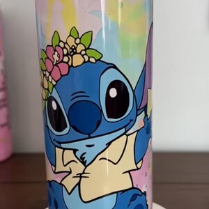 Op de afbeelding: Een hoge, cilindrische beker met een kleurrijk ontwerp van het personage Stitch uit Lilo & Stitch. Het blauwe personage is versierd met een bloemenkrans en staat tegen een aquarel-achtige achtergrond met roze, gele en blauwe tinten.