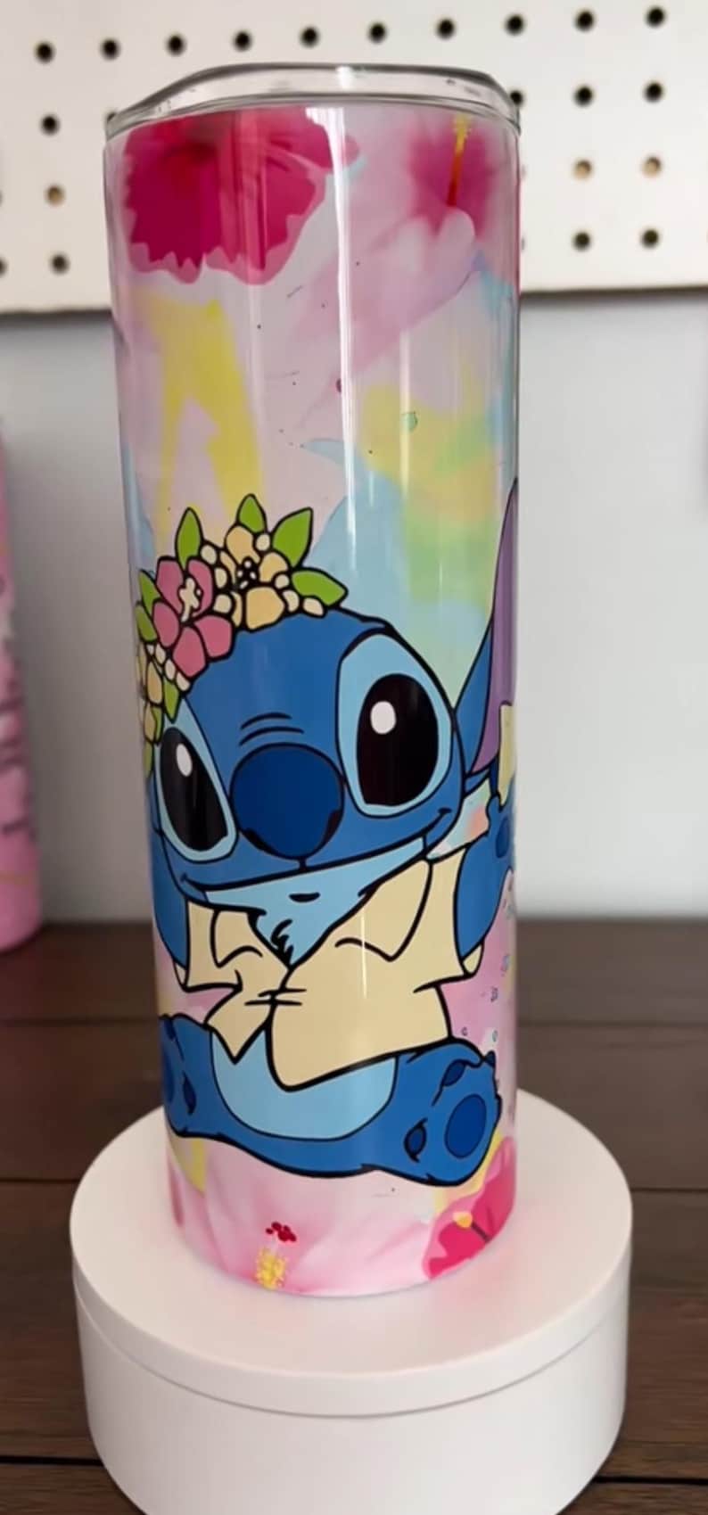 Op de afbeelding: Een hoge, cilindrische beker met een kleurrijk ontwerp van het personage Stitch uit Lilo & Stitch. Het blauwe personage is versierd met een bloemenkrans en staat tegen een aquarel-achtige achtergrond met roze, gele en blauwe tinten.
