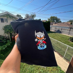 May include: Black hooded sweatshirt with an embroidered cartoon design of a superhero character. The design features a helmet, hammer, and cape in shades of white, red, blue, and yellow. The sweatshirt is held up in front of a green lawn.