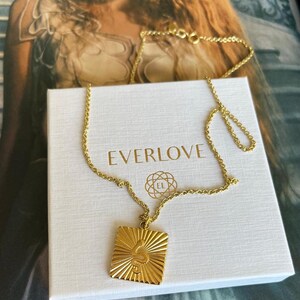 Puede incluir: Un collar de cadena dorada con un colgante cuadrado que presenta un diseño de serpiente estilizado. El colgante tiene un patrón de rayos de sol y el texto "EVERLOVE" es visible en la caja.