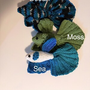 May include: Crocheted betta fish in shades of blue, green, and white. The fish are labeled "Ocean", "Moss", and "Sea". The fish have detailed fins and tails, and are displayed on a white surface.