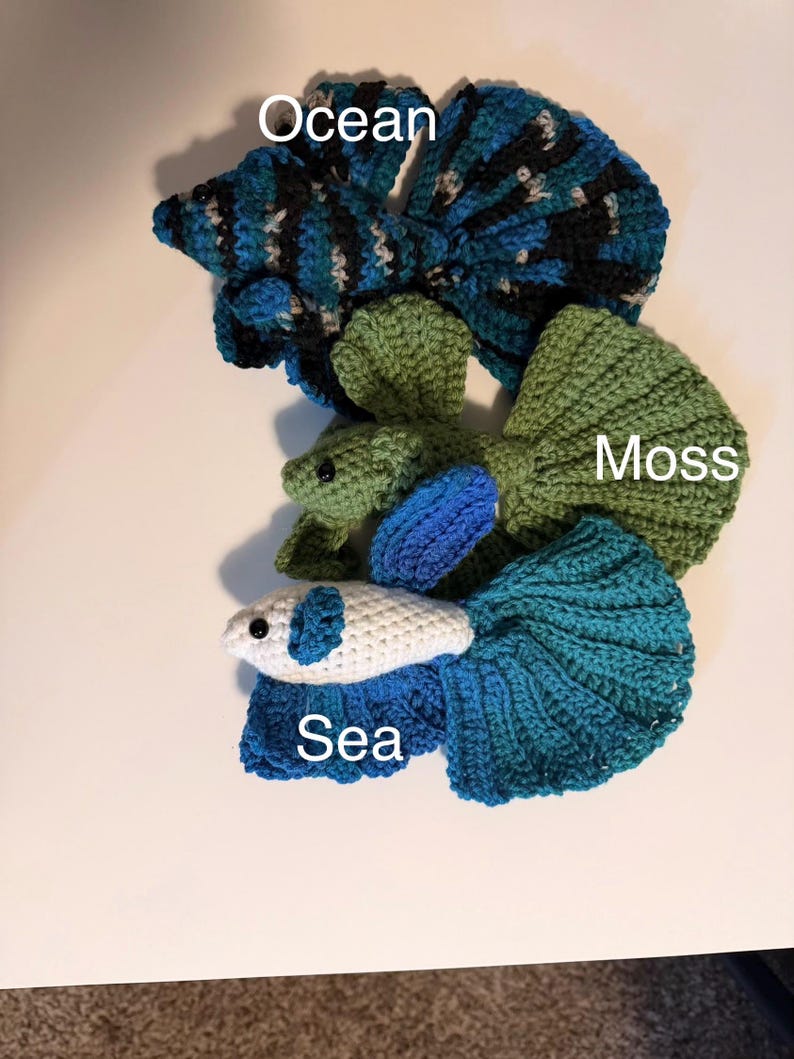 May include: Crocheted betta fish in shades of blue, green, and white. The fish are labeled "Ocean", "Moss", and "Sea". The fish have detailed fins and tails, and are displayed on a white surface.