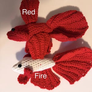 May include: Two handmade crochet fish. One is entirely red with the word "Red" above it. The other is white with red fins and tail, with the word "Fire" above it. Both have black bead eyes.