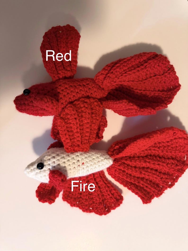May include: Two handmade crochet fish. One is entirely red with the word "Red" above it. The other is white with red fins and tail, with the word "Fire" above it. Both have black bead eyes.