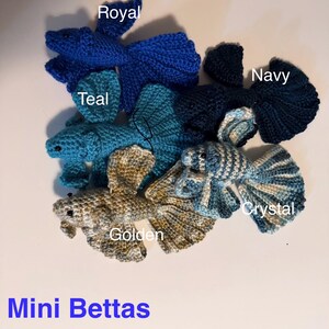 May include: A collection of handmade crochet betta fish in various colors, including royal blue, teal, navy, golden, and crystal. The text "Mini Bettas" is displayed in blue at the bottom of the image. These miniature fish are crafted with detailed fins and bodies.