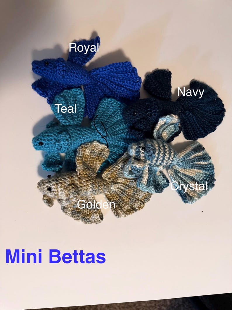 May include: A collection of handmade crochet betta fish in various colors, including royal blue, teal, navy, golden, and crystal. The text "Mini Bettas" is displayed in blue at the bottom of the image. These miniature fish are crafted with detailed fins and bodies.
