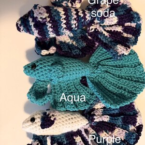 May include: Crocheted betta fish in three colors: "Grape soda" in blue, purple, and white; "Aqua" in teal; and "Purple" in white, blue, and purple. Each fish has a detailed tail and fins, with the color names written on the image.