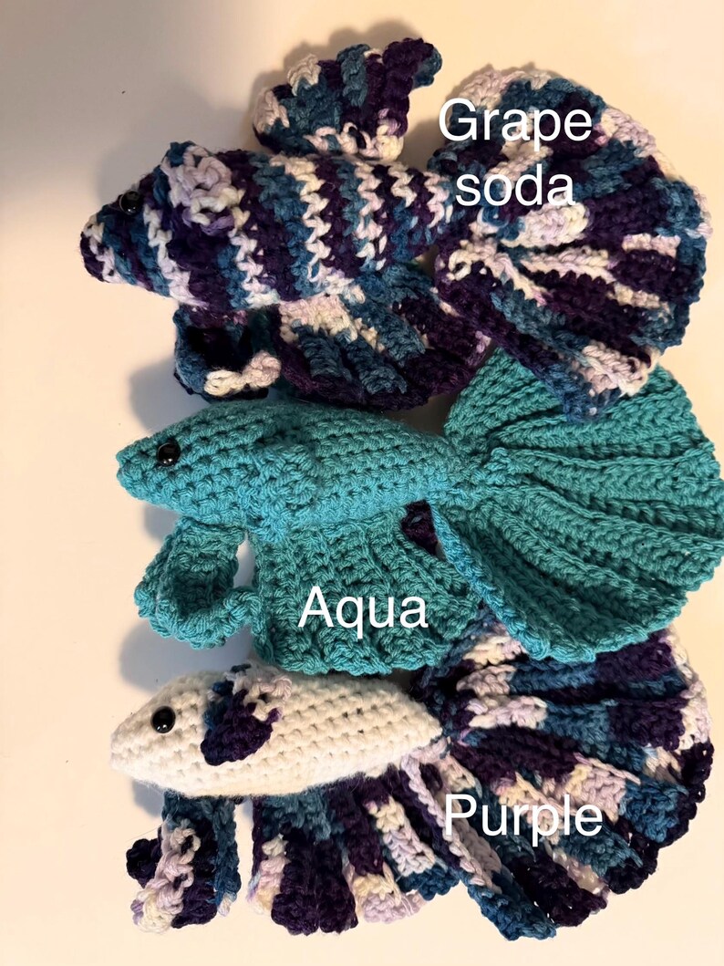 May include: Crocheted betta fish in three colors: "Grape soda" in blue, purple, and white; "Aqua" in teal; and "Purple" in white, blue, and purple. Each fish has a detailed tail and fins, with the color names written on the image.