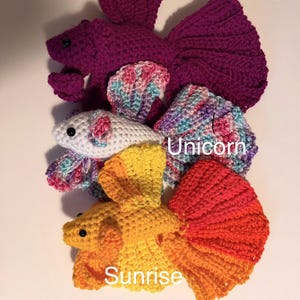May include: Crocheted betta fish in various colors, including purple, white, yellow, and orange. The fish are handmade and feature detailed fins and bodies. The text "Orchid", "Unicorn", and "Sunrise" are visible on the image.