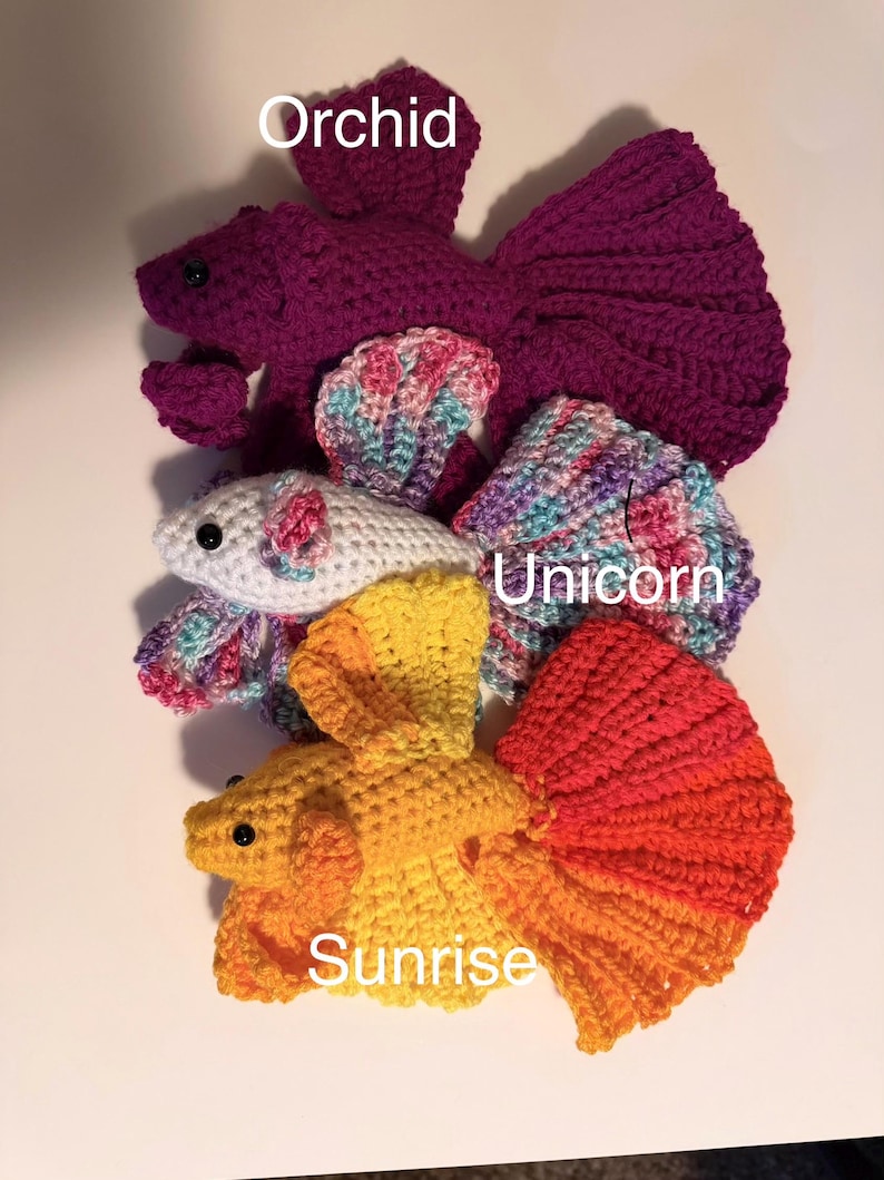 May include: Crocheted betta fish in various colors, including purple, white, yellow, and orange. The fish are handmade and feature detailed fins and bodies. The text "Orchid", "Unicorn", and "Sunrise" are visible on the image.
