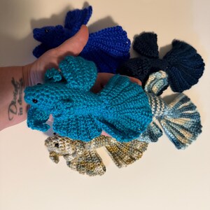 May include: A collection of handmade crochet fish in various shades of blue and a neutral color. The fish have detailed fins and tails, with small black eyes. The fish are displayed against a white background.