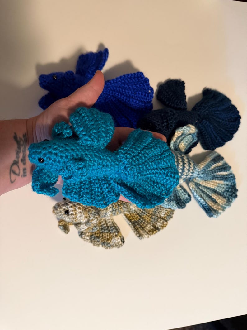 May include: A collection of handmade crochet fish in various shades of blue and a neutral color. The fish have detailed fins and tails, with small black eyes. The fish are displayed against a white background.