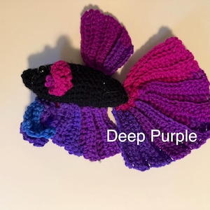 May include: A handmade crochet betta fish in shades of purple, magenta, and blue. The fish has a black head with a pink flower accent and the text "Deep Purple". This decorative item is a unique handmade gift.